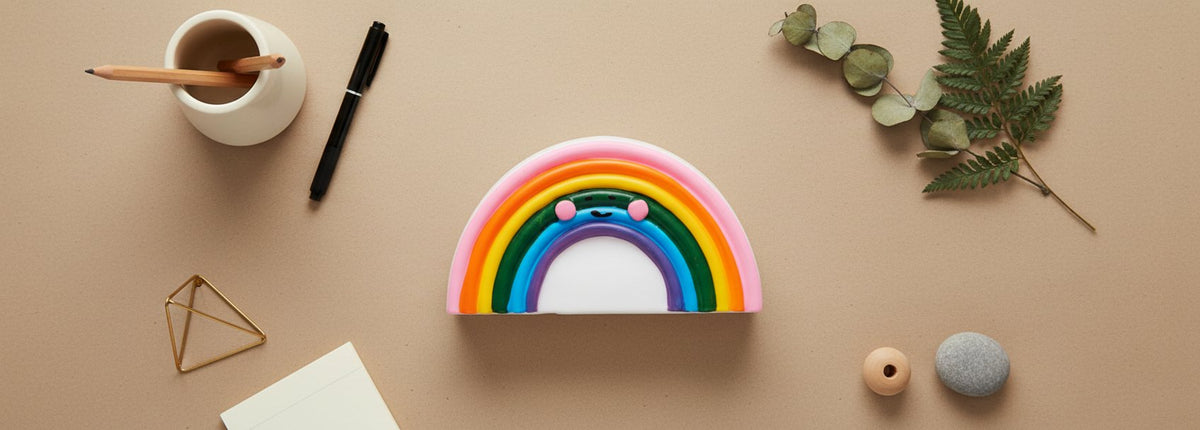 Lampada punto luce rainbow multicolore su scaffale, circondata da matite, penna, piante e oggetti decorativi su sfondo beige