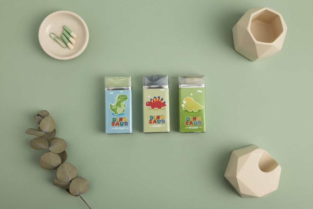 Gomme dinosauro colorate blu, beige e verde con packaging Dino Saur, circondate da accessori in ceramica e rami secchi su