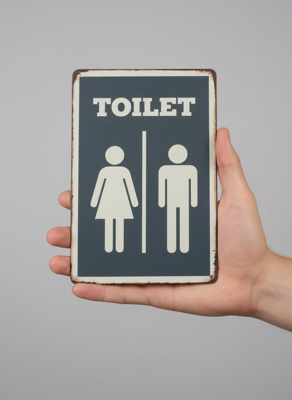 Placca toilet vintage in metallo grigio con figure stilizzate uomo e donna, bordi invecchiati, tenuta in mano su sfondo