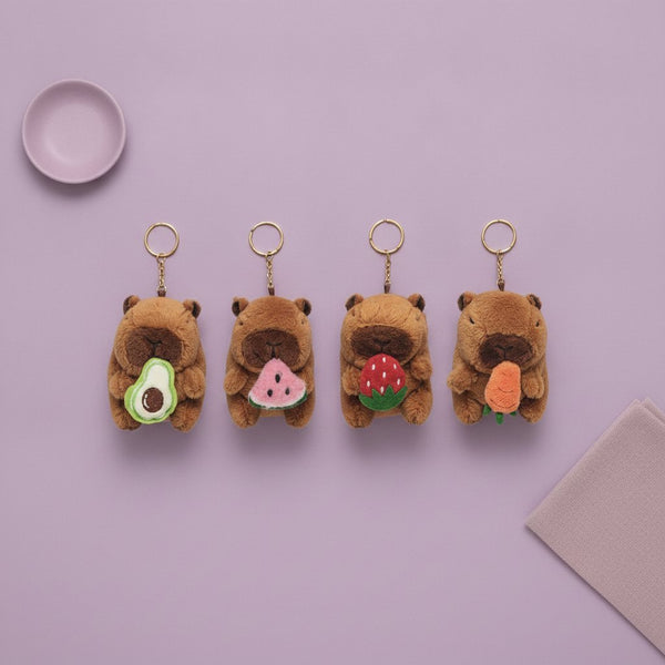 Quattro portachiavi in peluche a forma di capibara marrone con anellino dorato, ognuno con cibo diverso in bocca: avocado ver