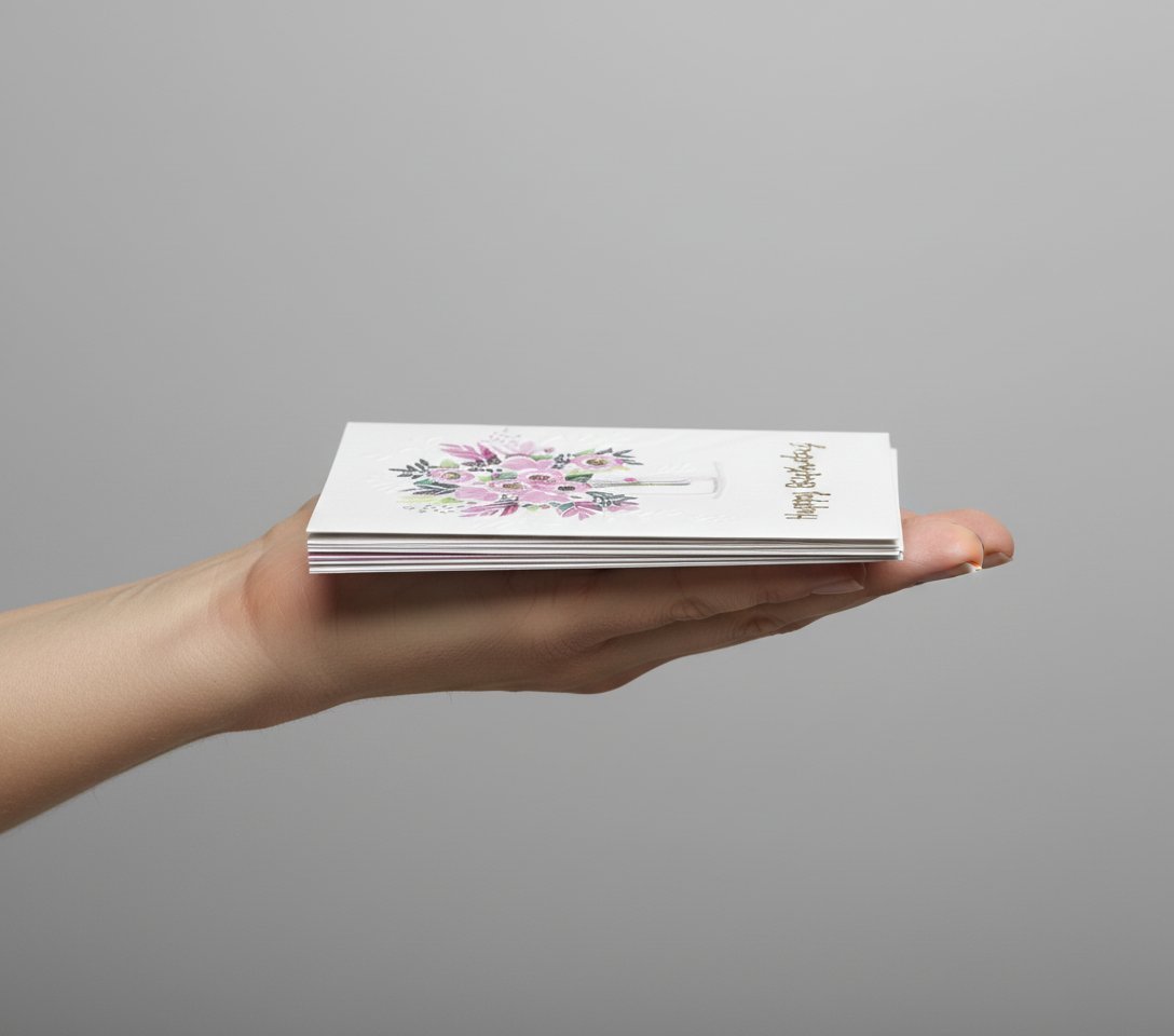 Biglietto auguri con bouquet di fiori rosa e viola, testo dorato, tenuto in mano su sfondo grigio