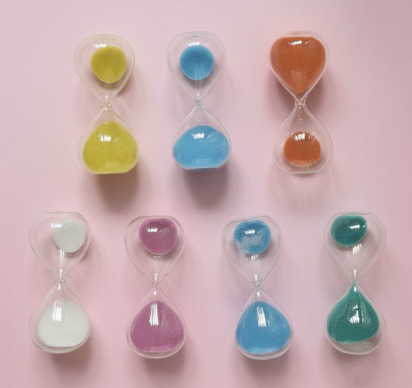 Sette clessidre colorate con sabbia gialla, blu, arancione, bianca, viola e verde su fondo rosa pastello